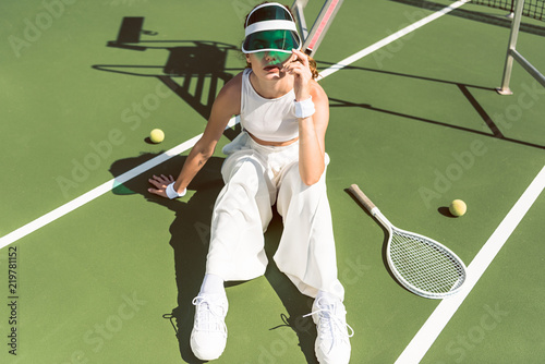 young woman in stylish white clothing and cap sitting on tennis court with racket and balls © LIGHTFIELD STUDIOS