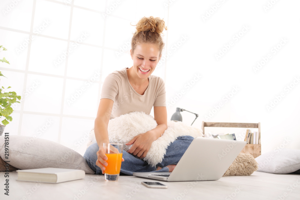 Fototapeta premium smiling young woman with computer drink orange juice,, sitting on the floor in living room on white wide window in the background