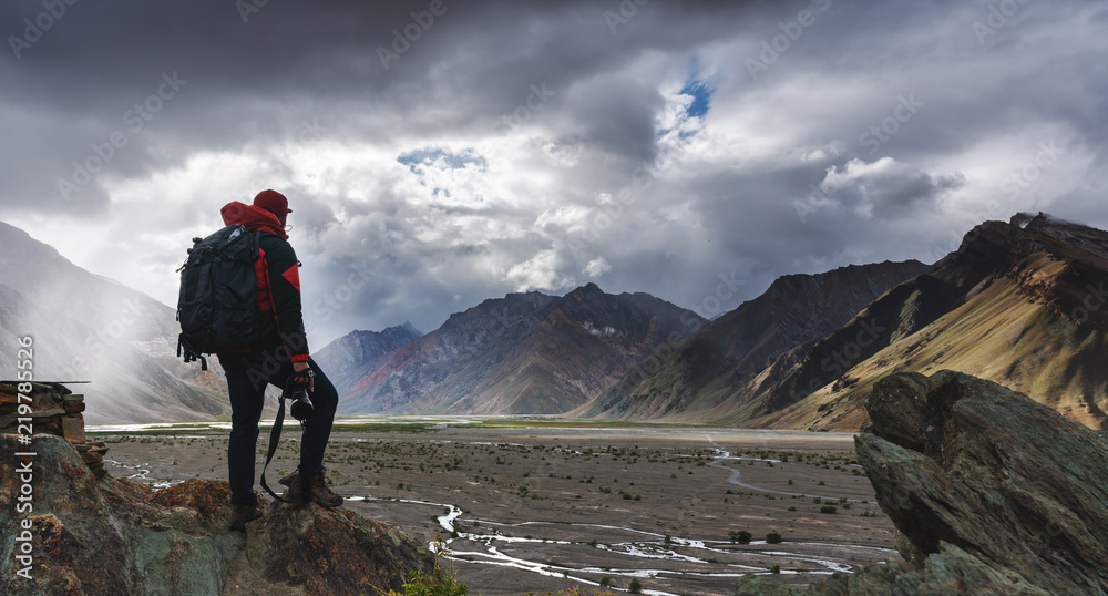 a man with backpack holding camera standing on cliff with mountains view and sunlight through cloud. Travel lifestyle, adventure and success concept