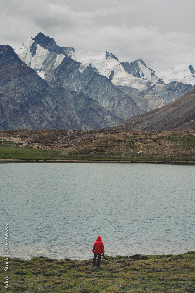 Traveller man in red jacket standing on meadow with lake and snow mountain view. Travel lifestyle, adventure and success  concept