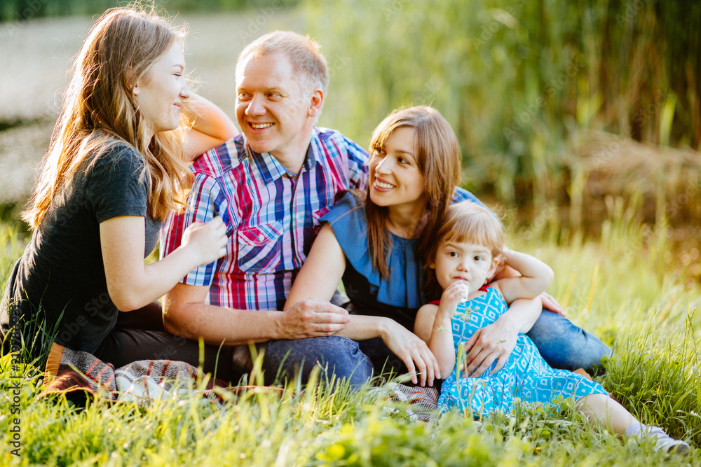 Happy family of four siting on the green grass at park in summer time. Cute teenager daughter spaeking about something while parent looking on her. Mother hugging little baby girl. Focus on teen girl.