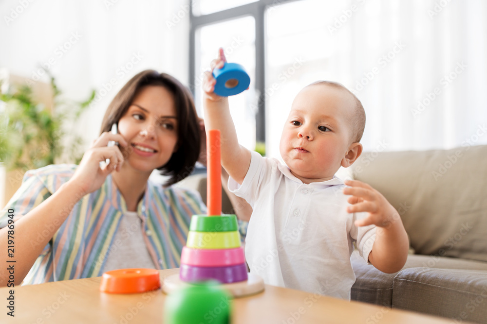 family, child and motherhood concept - happy baby boy playing developmental toy and mother calling on smartphone at home family, child and motherhood concept - happy baby boy playing developmental toy and mother calling on smartphone at home