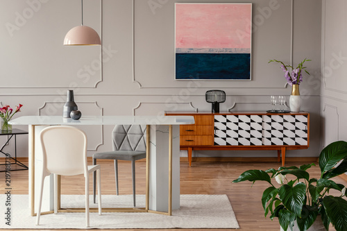 White chair and marble table under pink lamp in eclectic living room interior with painting above cabinet. Real photo photo