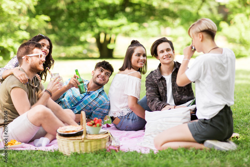 friendship, leisure and technology concept - group of happy smiling friends with non alcoholic drinks photographing at picnic in summer park