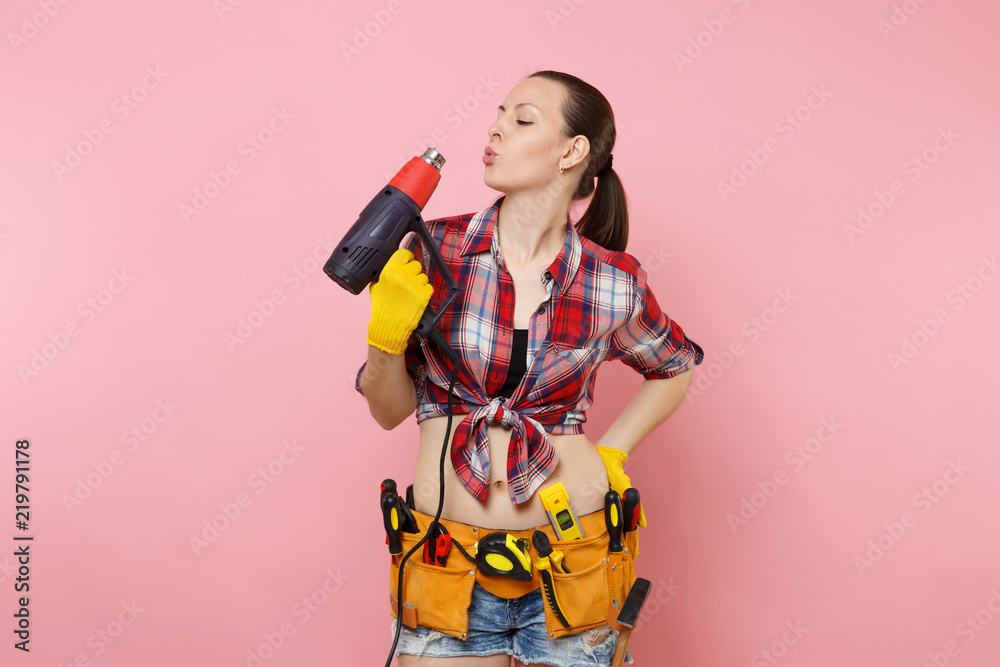 Young beautiful fun handyman woman in plaid shirt, denim shorts, kit tools belt full of variety instruments holding power heat gun isolated on pink background. Female in male work. Renovation concept. Young beautiful fun handyman woman in plaid shirt, denim shorts, kit tools belt full of variety instruments holding power heat gun isolated on pink background. Female in male work. Renovation concept.
