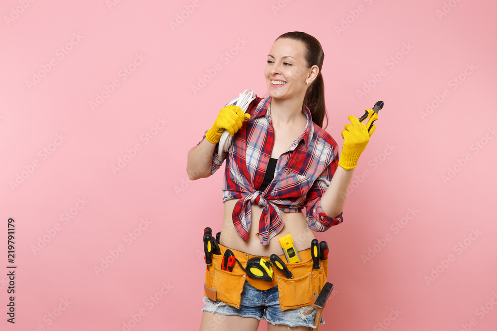 Young excited handyman woman in plaid shirt, denim shorts, kit tools belt full of variety instruments holding electric power cabel isolated on pink background. Female in male work. Renovation concept Young excited handyman woman in plaid shirt, denim shorts, kit tools belt full of variety instruments holding electric power cabel isolated on pink background. Female in male work. Renovation concept