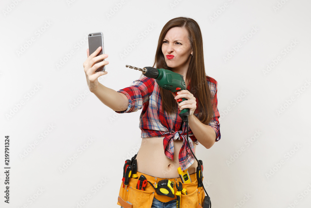 Strong excited handyman woman in shirt, kit tools belt full of instruments  doing selfie on mobile phone with power electric drill isolated on white  background. Female in male work. Renovation concept. Photos