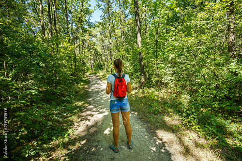 Young girl backpacker in jeans shorts with red backpack and sandals on his feet  walks on the road in the green forest in the sunshine outdoors in the summer time
