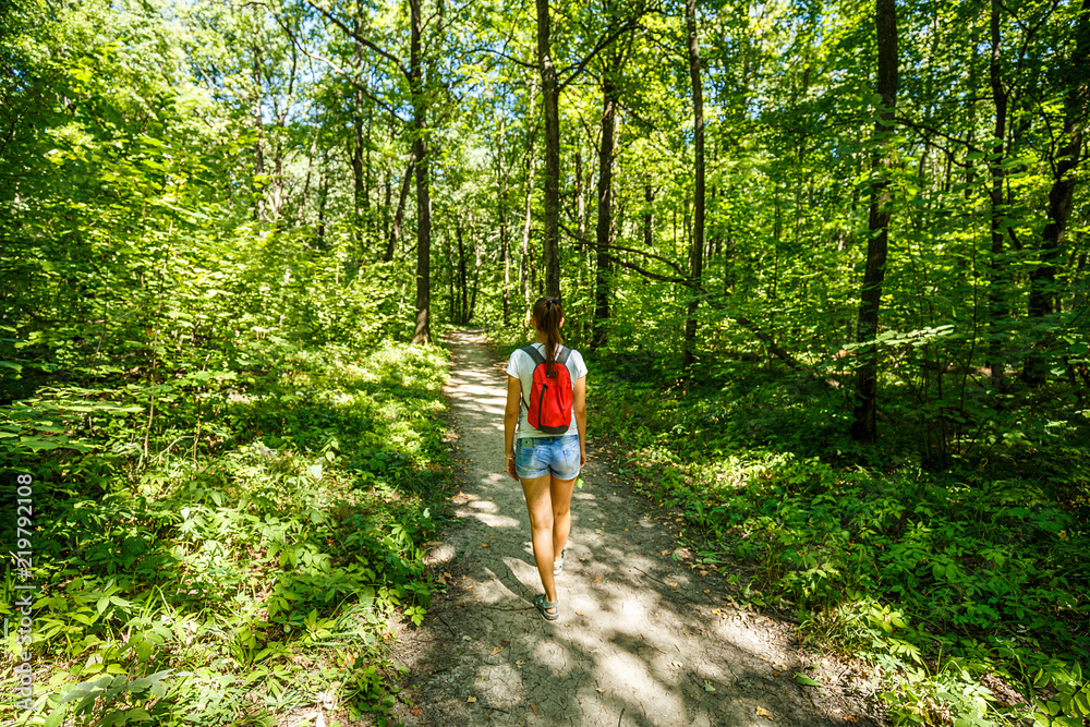 Young girl backpacker in jeans shorts with red backpack and sandals on his feet, walks on the road in the green forest in the sunshine outdoors in the summer time