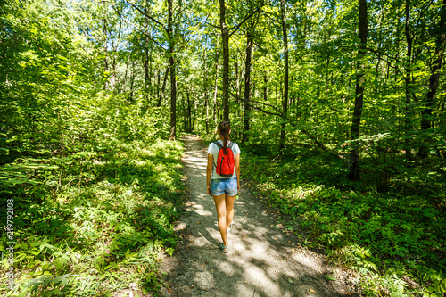 Young girl backpacker in jeans shorts with red backpack and sandals on his feet  walks on the road in the green forest in the sunshine outdoors in the summer time