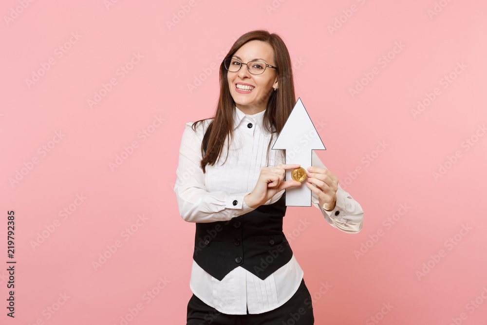 Fototapeta premium Young joyful attractive business woman in black suit, glasses holding bitcoin, up arrow isolated on pastel pink background. Lady boss. Achievement career wealth concept. Copy space for advertisement.