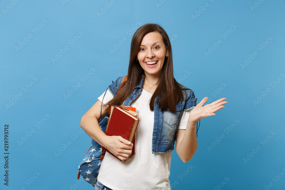 Portrait of young surprised lovely woman student with backpack pointing hand aside holding school books ready to learn isolated on blue background. Education in high school university college concept.