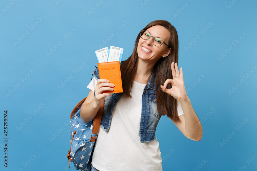 Young joyful woman student in glasses with backpack holding passport, boarding pass tickets and showing OK sign isolated on blue background. Education in university college abroad. Air travel flight. Young joyful woman student in glasses with backpack holding passport, boarding pass tickets and showing OK sign isolated on blue background. Education in university college abroad. Air travel flight.
