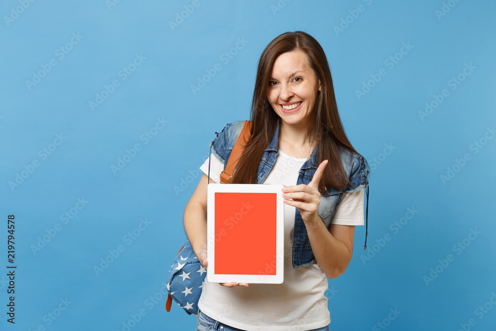 Obraz premium Young joyful pretty woman student pointing index finger up holding tablet pc computer with blank black empty screen isolated on blue background. Education in university. Copy space for advertisement.