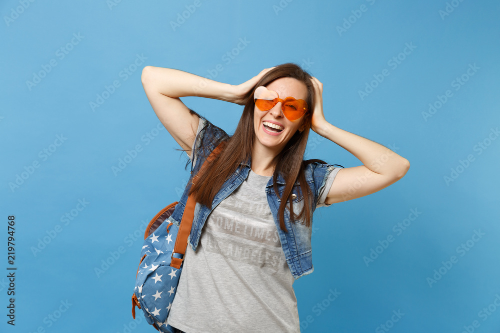 Portrait of young laughing joyful beautiful woman student with backpack in orange heart glasses clinging to head isolated on blue background. Education in high school. Copy space for advertisement.