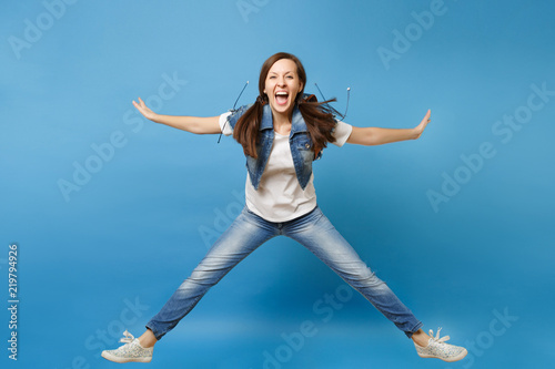 Full length portrait of young overjoyed woman student in denim clothes jumping spreading hands and legs screaming isolated on blue background. Education in high school. Copy space for advertisement.