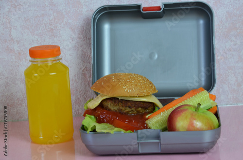 Homemade Hamburgers with lettuce in lunchbox on the table. with bright yellow orange juice. with banana nuts. Sliced vegetables carrots and cucumber. Opposite the school board. Snack. School lunch