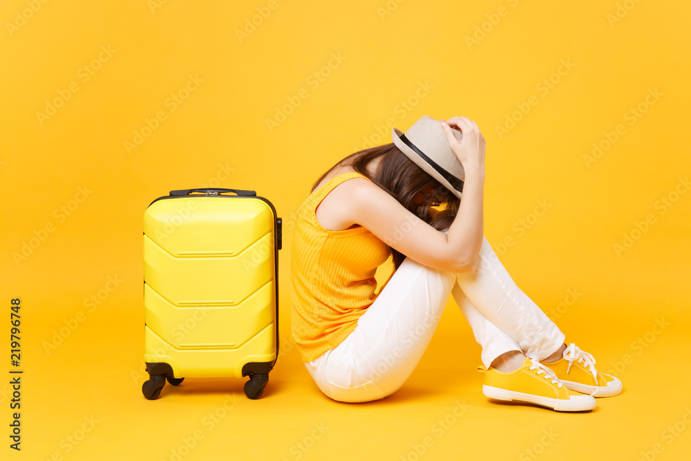 Sad traveler tourist woman in summer casual clothes, hat sit near suitcase  isolated on yellow orange background. Female passenger traveling abroad to  travel on weekends getaway. Air flight concept. Stock Photo
