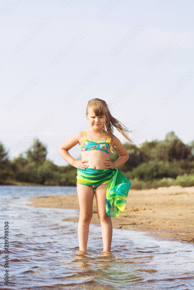 little joyful blonde with a tail girl in a swimsuit and a pareo posing and jumping on the river bank in sunny summer weather little joyful blonde with a tail girl in a swimsuit and a pareo posing and jumping on the river bank in sunny summer weather