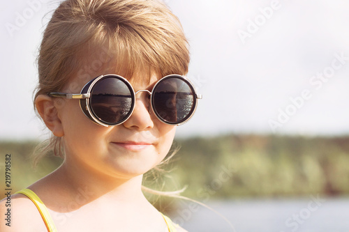 Portrait of a small blond girl with glasses on the river bank in sunny summer weather