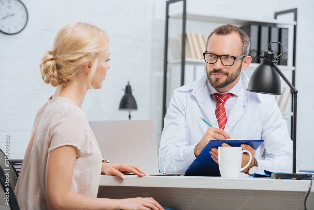 Obraz premium chiropractic in white coat making notes in notepad with female patient near by at table during appointment in clinic