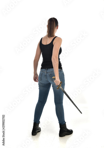 full length portrait of brunette girl wearing black single and jeans. standing pose holding a sword. isolated on white studio background.