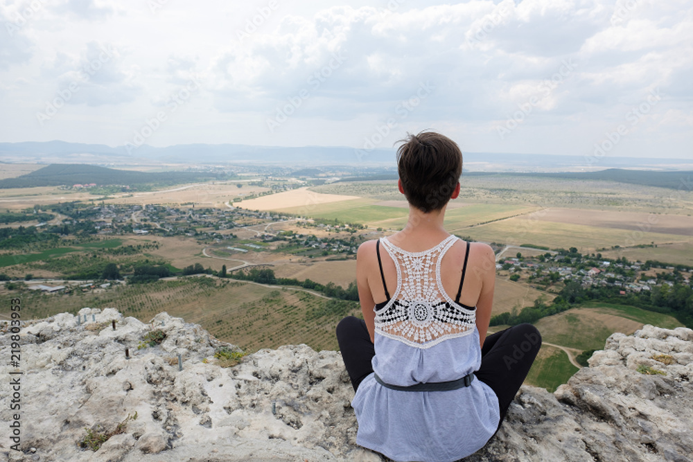 young woman sitting on peak of a cliff and looking forward to the plain. Female hiker  on top of mountain and enjoying view. Travel, freedom, vacation, trekking, hiking, zen concept