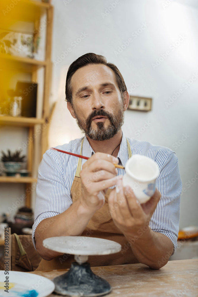 Bearded ceramist. Dark-eyed bearded ceramist wearing apron working in his light spacious workshop Bearded ceramist. Dark-eyed bearded ceramist wearing apron working in his light spacious workshop