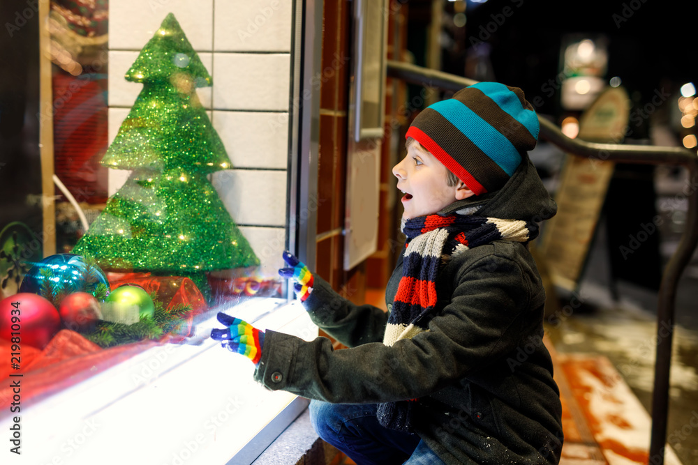 Cute little school kid boy on Christmas market. Funny happy child in fashion winter clothes making window shopping decorated with gifts, xmas tree. Snow falling down, snowfall:
