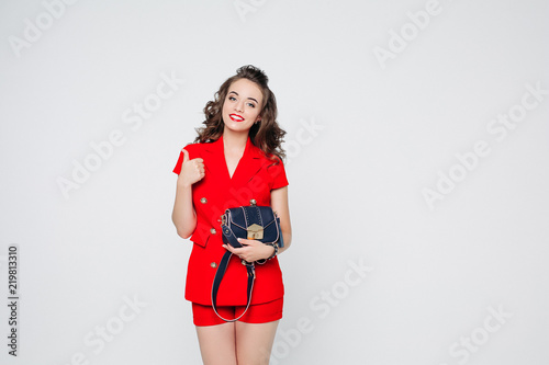 Studio portrait of attractive smiling woman with hairstyle and make up wearing bright red suit and holding trendy blue leather crossbag, showing thumb up with toothy smile. Isolated on white. photo