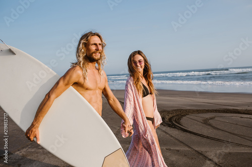 happy surfer walking on beach with girlfriend and holding surfing board in bali, indonesia