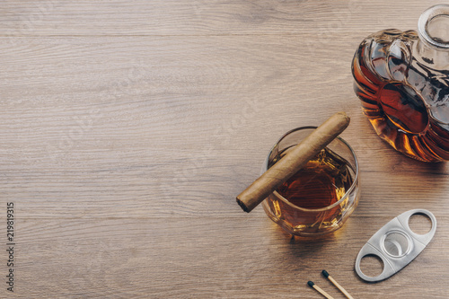 Cuban cigar on a glass of bourbon whiskey, a whiskey decanter with a stainless steel cigar cutter and matches on the wooden table