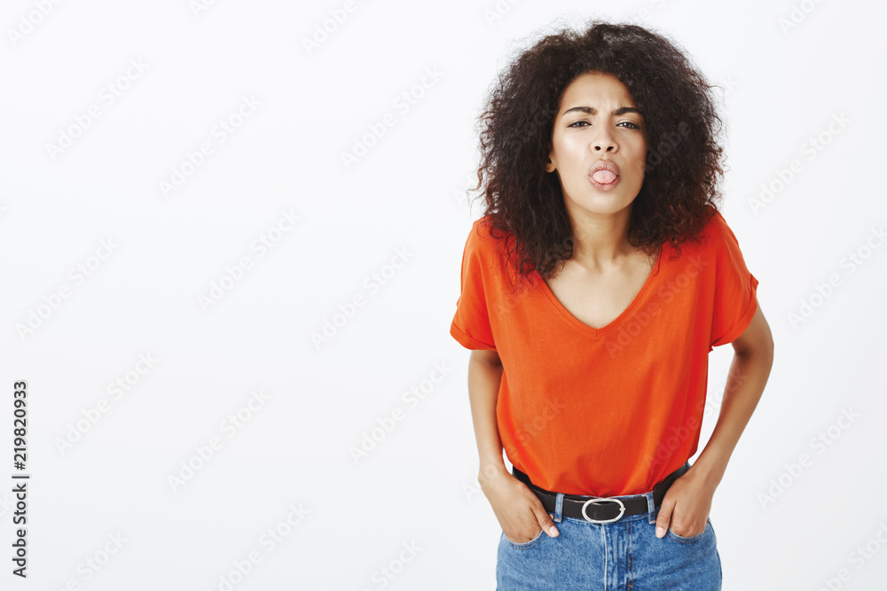 Girl with childish behaviour having quarrel. Displeased funny dark-skinned female student with afro hairstyle, holding hands in pockets, bending towards camera and sticking out tongue while frowning