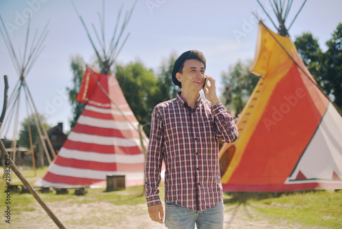 Young man tourist talking on the phone and smiling on the background teepee / tipi- native indian house. Man in hat traveler in sunny day.