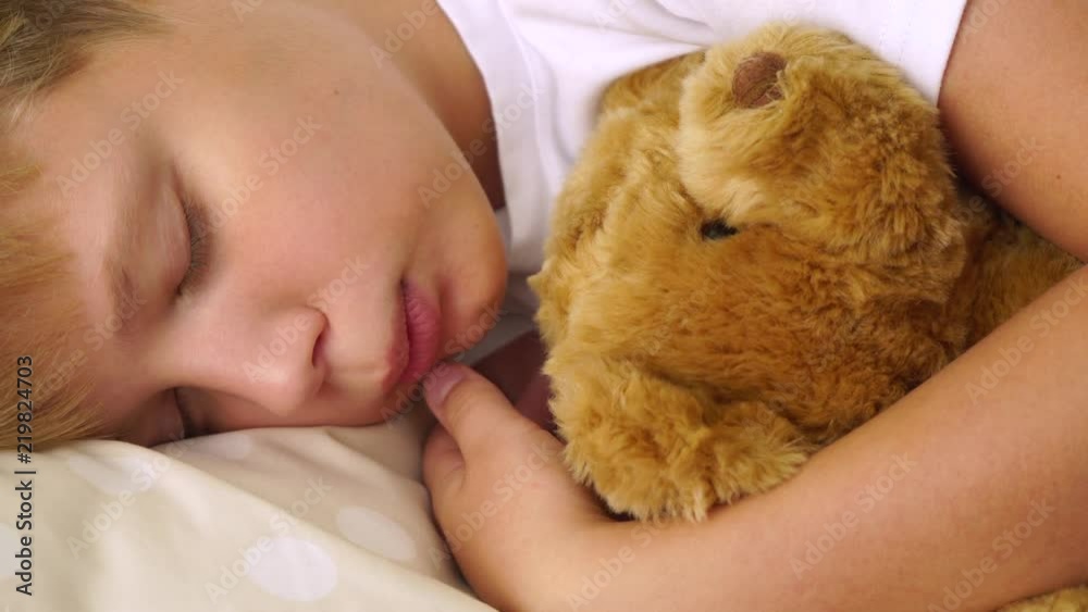 Closeup portrait of cute sleeping calm young white kid lying in his comfortable bed hugging cute brown soft teddy bear toy. Real time 4k video footage.