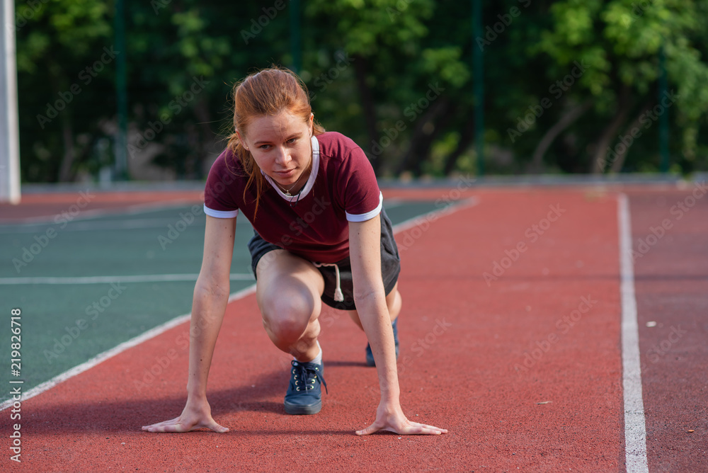 the red-haired student in shorts gives the standards for running for a short distance at the physical education class at an open sports ground.Female athlete ready to run on running track