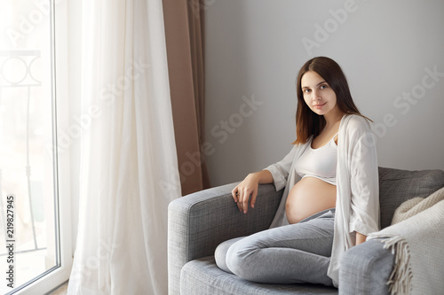 Husband taking picture of his charming pregnant wife sitting near window on armchair, smiling with cute and tender expression at camera, being calm and happy while waiting for daughter to be born