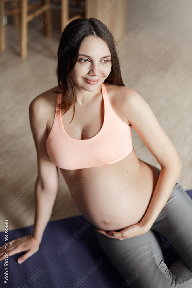 Vertical shot of cute happy european young pregnant woman in sports bra and  leggings, leaning on
