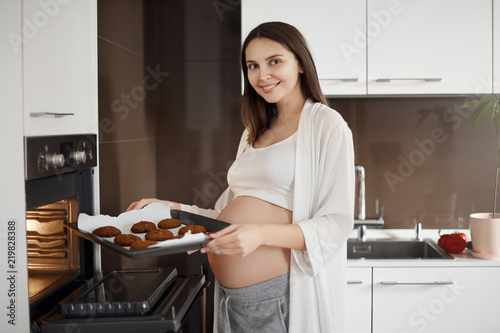 Cute and happy pregnant woman likes to eat sweet, baking at home while resting during pregnancy and having nothing to do. Charming mother taking cookies out of oven and holding dripping-pan