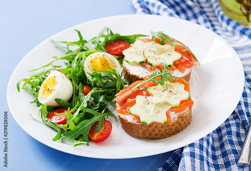 Tasty breakfast. Open sandwiches with salmon, cream cheese and rye bread in a white plate and salad with tomato, egg and arugula.