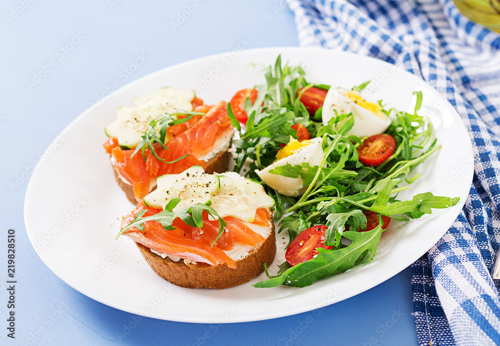 Tasty breakfast. Open sandwiches with salmon, cream cheese and rye bread in a white plate and salad with tomato, egg and arugula.