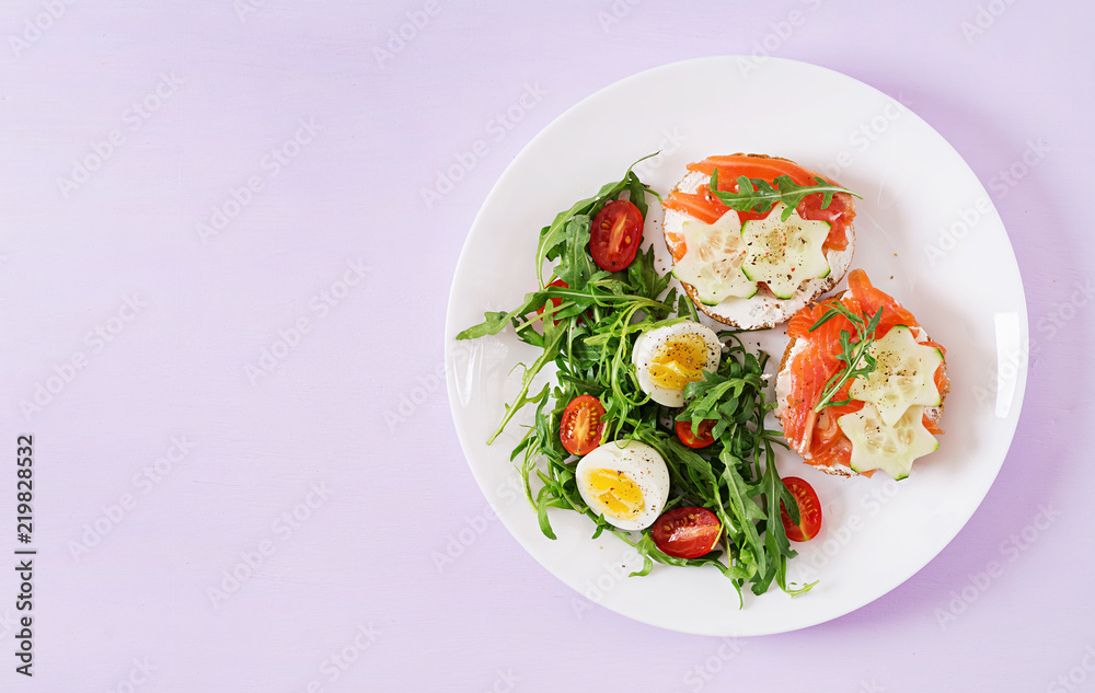 Tasty breakfast. Open sandwiches with salmon, cream cheese and rye bread in a white plate and salad with tomato, egg and arugula. Top view. Flat lay Tasty breakfast. Open sandwiches with salmon, cream cheese and rye bread in a white plate and salad with tomato, egg and arugula. Top view. Flat lay