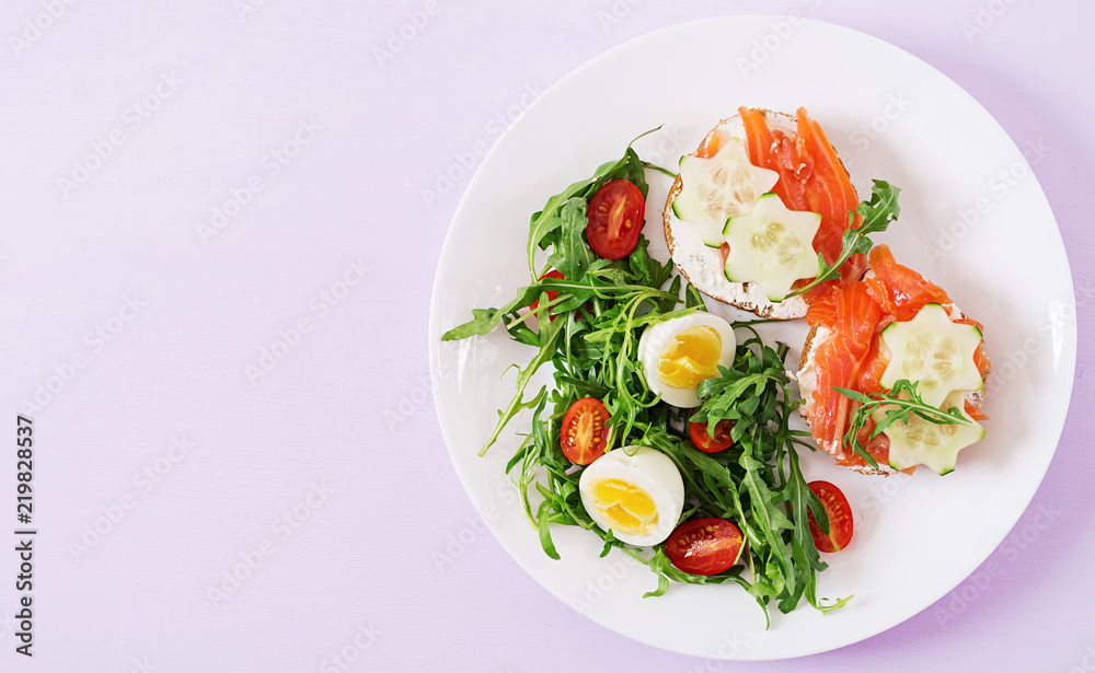 Tasty breakfast. Open sandwiches with salmon, cream cheese and rye bread in a white plate and salad with tomato, egg and arugula. Top view. Flat lay Tasty breakfast. Open sandwiches with salmon, cream cheese and rye bread in a white plate and salad with tomato, egg and arugula. Top view. Flat lay