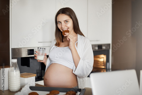 Pregnant woman do not share food. Charming and caring european wife with cute belly  standing near kitchen table with cookies on dripping-pan  biting cookies joyfully and holding glass of milk