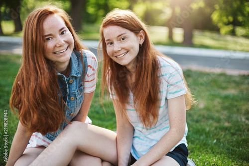 Close-up shot of beautiful european sisters with red hair and freckles sitting on green grass and smiling broadly at camera, hanging out with friends on picnic, expressing joy and amusement