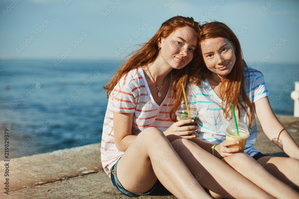 Let troubles go away when you near. Charming female siblings with ginger hair and freckles sitting near seashore and enjoying sunny day, drinking cocktails, girl leaning on sister shoulder and smiling