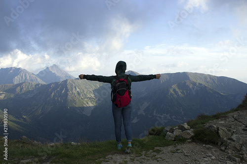 Young female tourist standing with her arms wide open and looks up, mountains on background . Back view