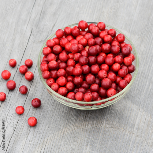 The ripe red berries (Vaccinium vitis-idaea) are in a transparent glass bowl on a wooden texture. The collected wild cowberry (foxberry, lingonberry) is in outdoor.