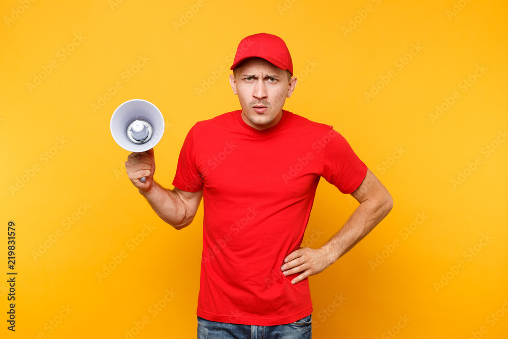 Delivery service man in red uniform isolated on yellow background. Male employee courier in cap, t-shirt screaming in megaphone hot news. Fun guy announces discounts sale. Copy space for advertisement