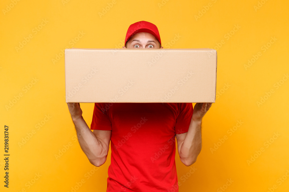 Delivery man in red uniform isolated on yellow orange background. Professional male employee courier or dealer in cap t-shirt holding empty cardboard box. Receiving package service concept. Copy space Delivery man in red uniform isolated on yellow orange background. Professional male employee courier or dealer in cap t-shirt holding empty cardboard box. Receiving package service concept. Copy space